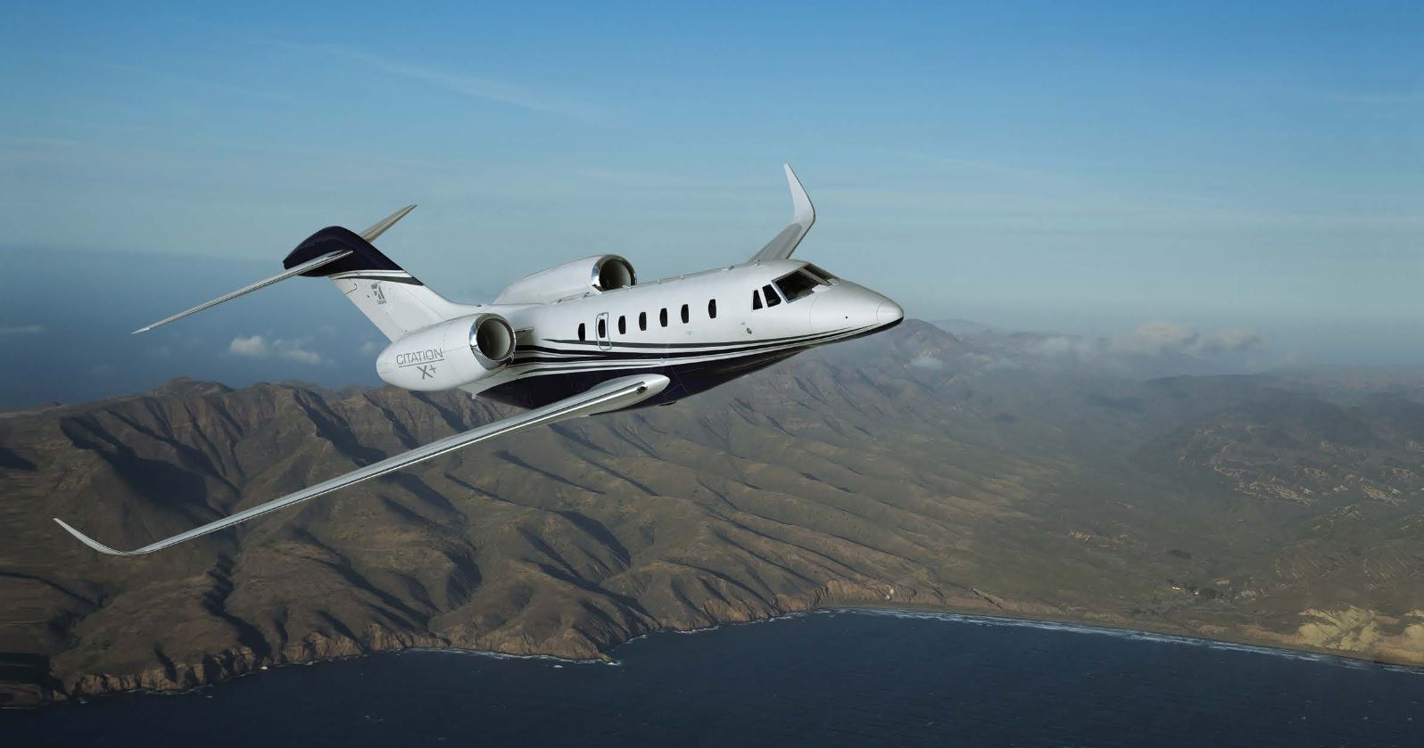 A Cessna Citation X+ flying above mountainous terrain.