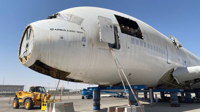 A close-up, low-angle view of the massive, weathered white fuselage of an Airbus A380-800, which is being dismantled at an industrial facility. The large aircraft body is elevated high off the ground, supported by several cylindrical blue metal jacks. Its nose cone is missing, and the cockpit windows are covered with silver tape. A yellow construction vehicle is parked near the front of the aircraft on the tarmac under a bright blue sky.