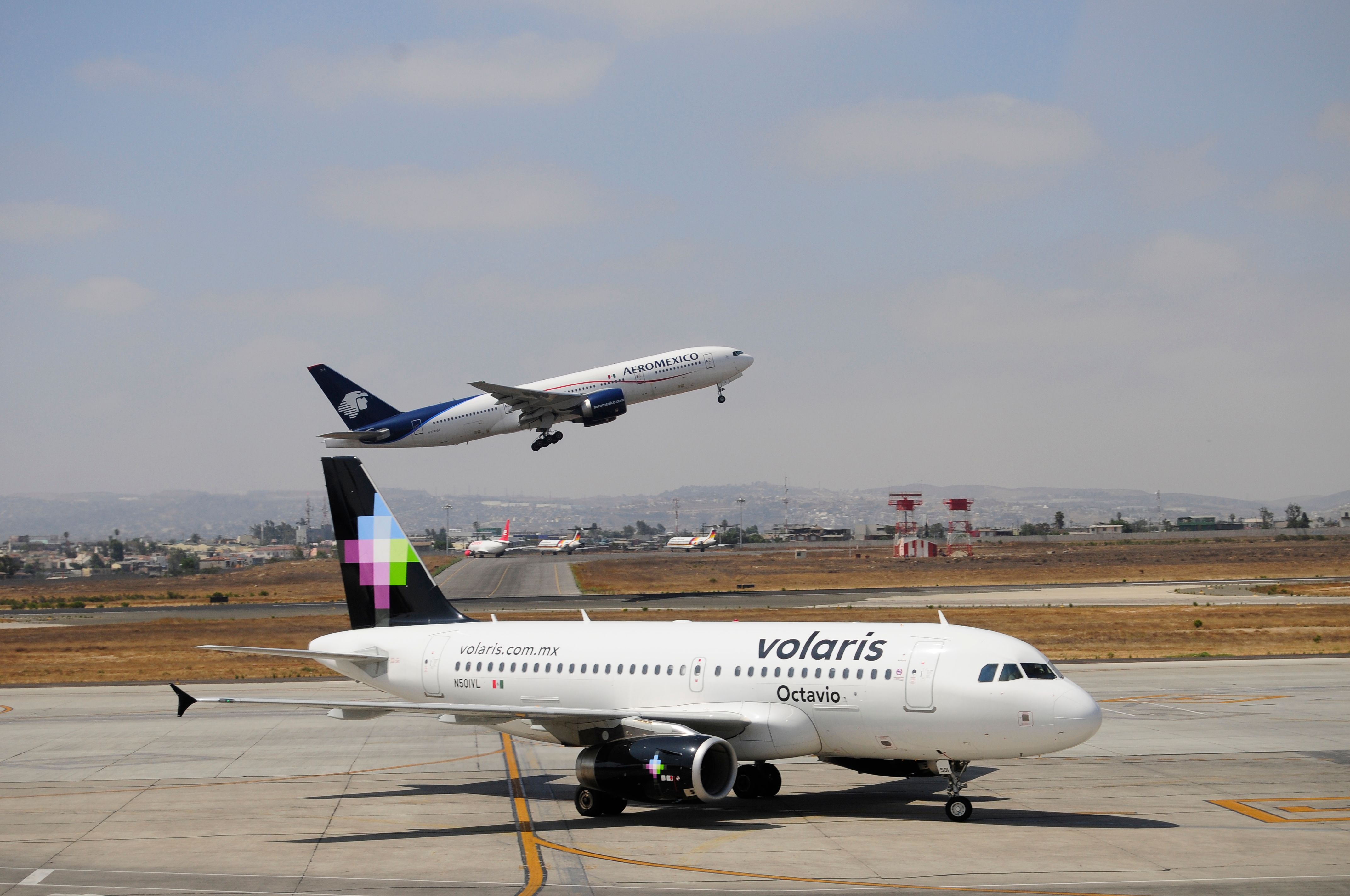 A Volaris aircraft on an airport apron as an Aeromexico aircraft takes off.