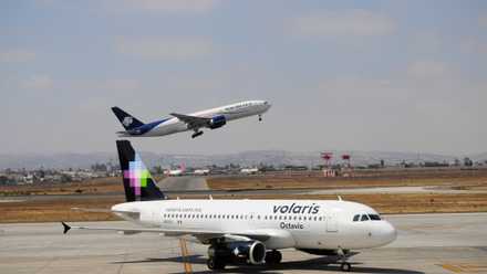 A Volaris aircraft on an airport apron as an Aeromexico aircraft takes off.