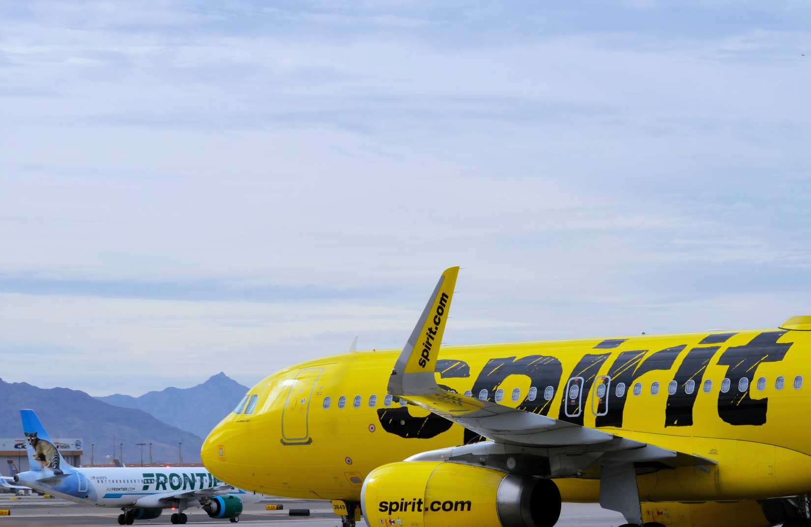 A bright yellow Spirit Airlines Airbus A320 passenger jet departs from Phoenix Sky Harbor International Airport (PHX), with a Frontier Airlines aircraft in the background