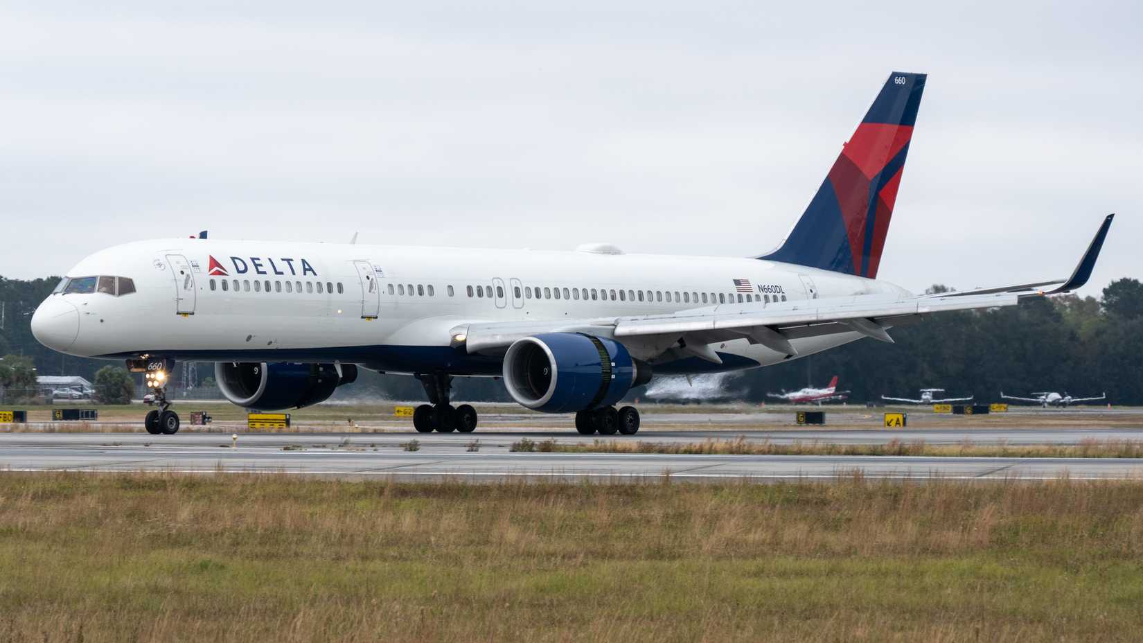 A Delta Air Lines Boeing 757 aircraft taxiing on a runway at an airport, with a cloudy sky and trees in the background.