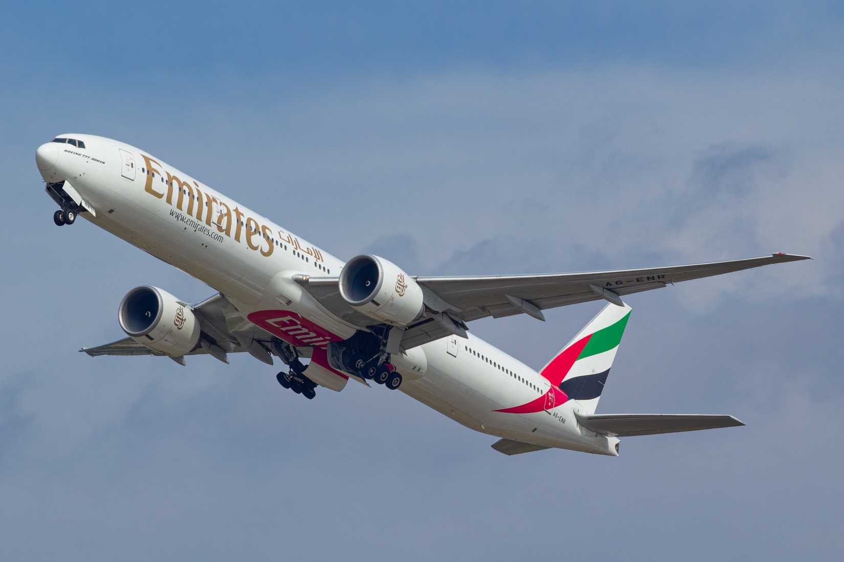 An Emirates Boeing 777-300ER (A6-ENR) in standard livery, with its gear still down as it climbs after takeoff against a partly cloudy sky.