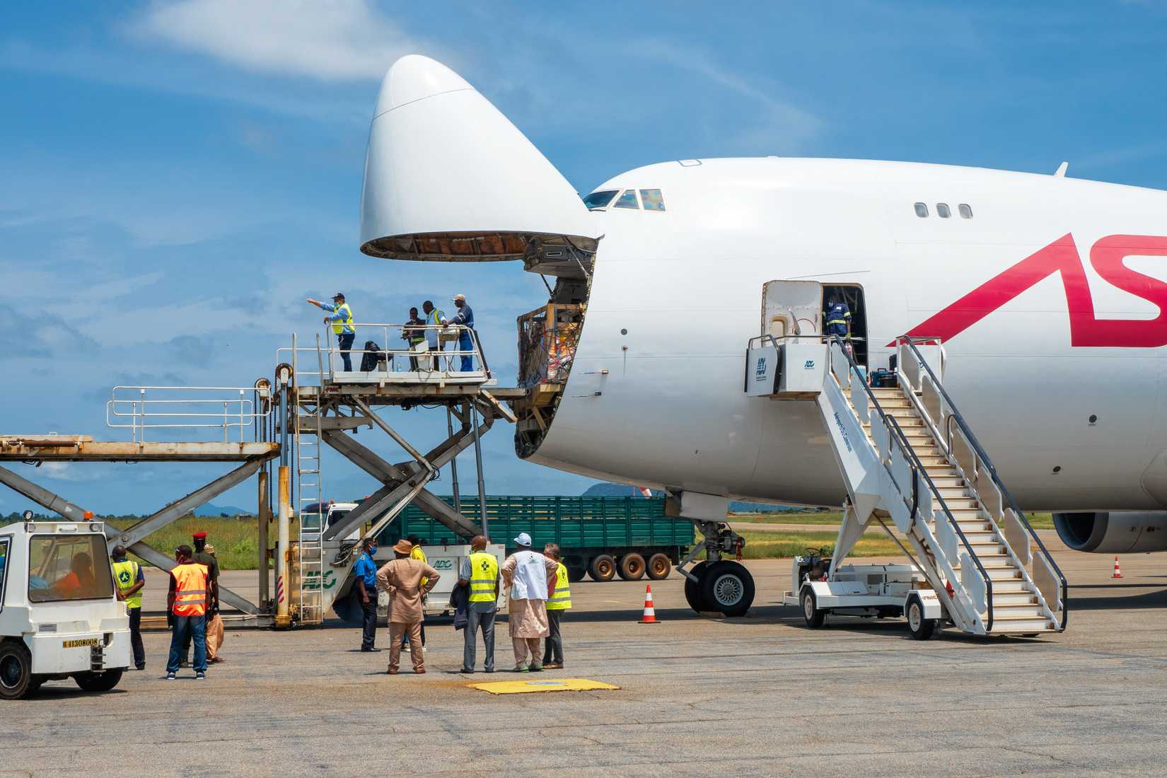 A Boeing 747 Jumbo Jet freighter aircraft with a wide open nose cargo door being offloaded by a high-loader at a cargo ramp