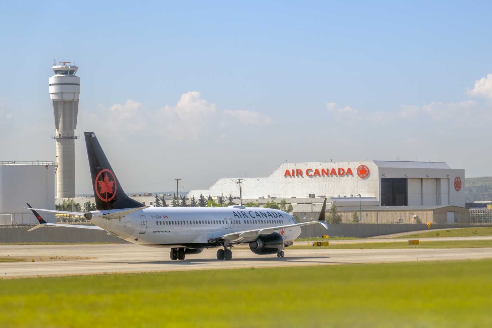 . Ein Boeing 737 MAX 8-Flugzeug von Air Canada rollt auf der Landebahn mit einem Flugsicherungsturm und einem Air Canada-Hangar im Hintergrund.
