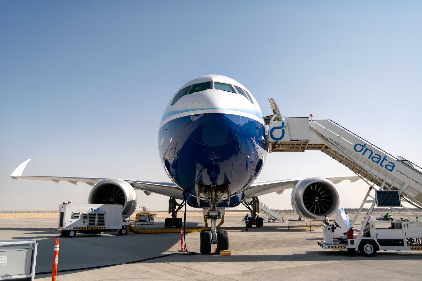 A low-angle, head-on view of a Boeing 777X jet with a distinctive dark blue and white livery, parked on a tarmac. A mobile passenger airstair branded "dnata" is connected to the main door on the right. Various ground power units and service vehicles are positioned around the aircraft's engines and fuselage. The scene takes place in a sunny, arid environment under a clear blue sky.