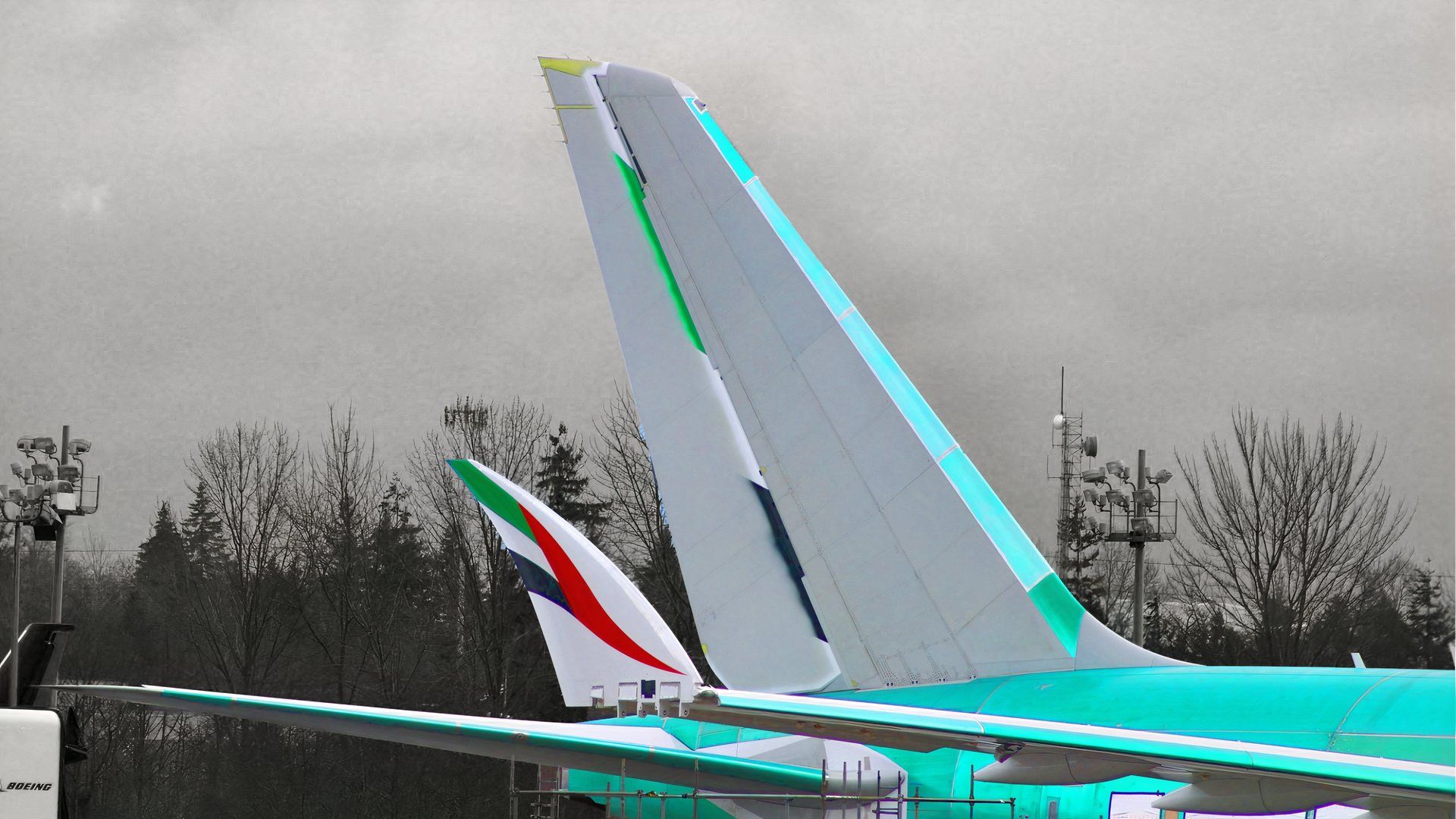 A close-up photograph of a single Boeing 777X aircraft parked on a tarmac. The aircraft's dominant vertical stabilizer is painted in a light gray with bright teal accents. The wingtip of the same aircraft, visible in the foreground to the left, features the red, green, and white UAE flag colors of the Emirates livery. The plane is parked against a backdrop of dark, bare trees and an overcast gray sky.