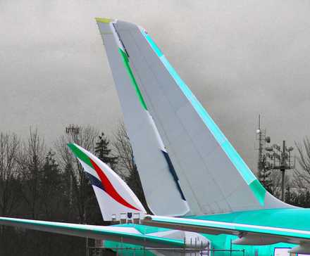 A close-up photograph of a single Boeing 777X aircraft parked on a tarmac. The aircraft's dominant vertical stabilizer is painted in a light gray with bright teal accents. The wingtip of the same aircraft, visible in the foreground to the left, features the red, green, and white UAE flag colors of the Emirates livery. The plane is parked against a backdrop of dark, bare trees and an overcast gray sky.