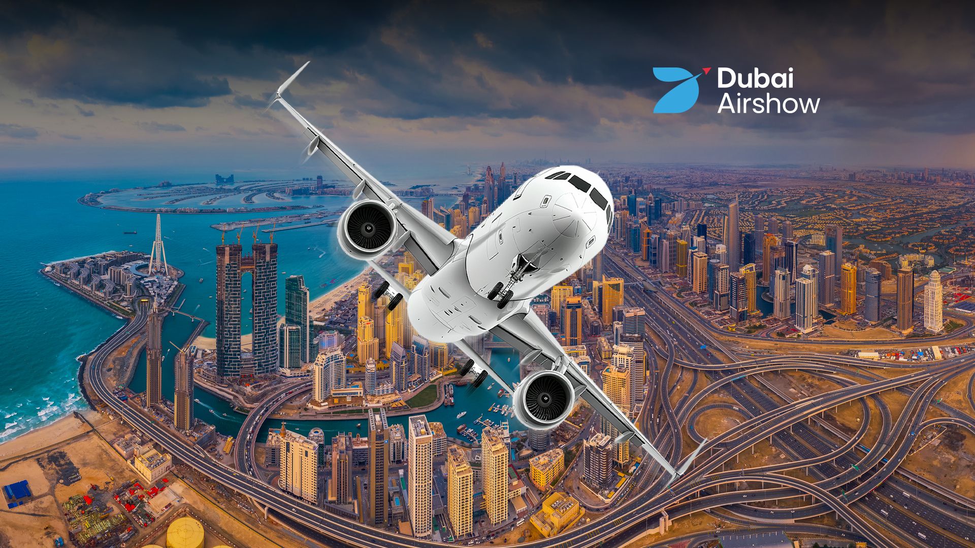 A promotional image for the Dubai Airshow, featuring a white passenger jet performing a steep bank over a dramatic, wide-angle aerial view of the Dubai skyline. Iconic landmarks like the Palm Jumeirah and the Burj Al Arab are visible along the coast, alongside a dense cityscape of skyscrapers and complex highway interchanges. The 