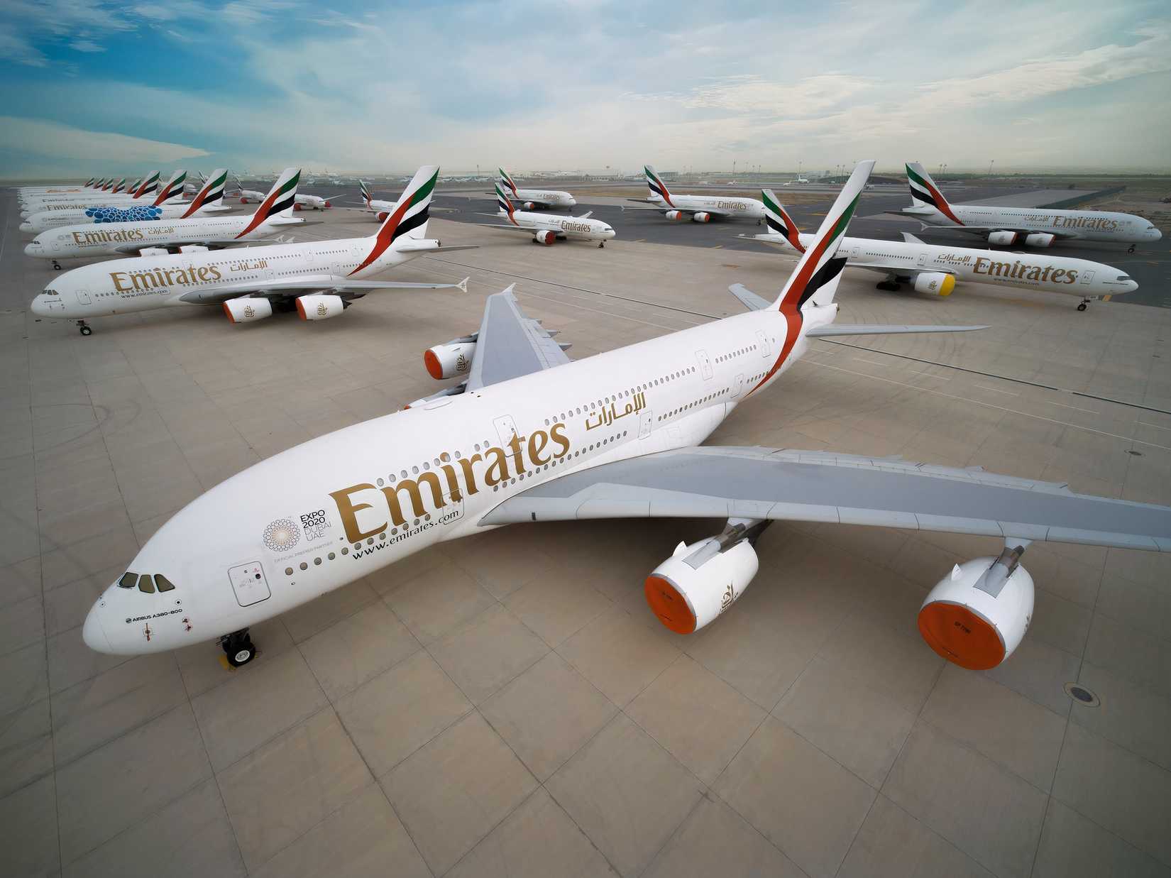 A high-angle photograph showing a large number of Emirates Airbus A380 aircraft parked closely together on a wide airport tarmac. The nearest aircraft features the gold "Emirates" livery and the UAE flag tail design, with orange protective covers on its engine inlets. A vast, uniform line of identical A380 tail fins extends across the background towards the horizon under a cloudy sky.
