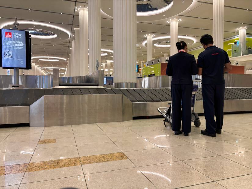 The interior of a vast, modern airport baggage claim hall, characterized by tall, white fluted columns and circular ceiling lights. In the foreground, two airport service staff members in dark uniforms stand with their backs to the camera, looking down at a mobile trolley. A large metallic baggage carousel dominates the middle ground, and a suspended digital screen on the left displays an Emirates Skywards promotion.