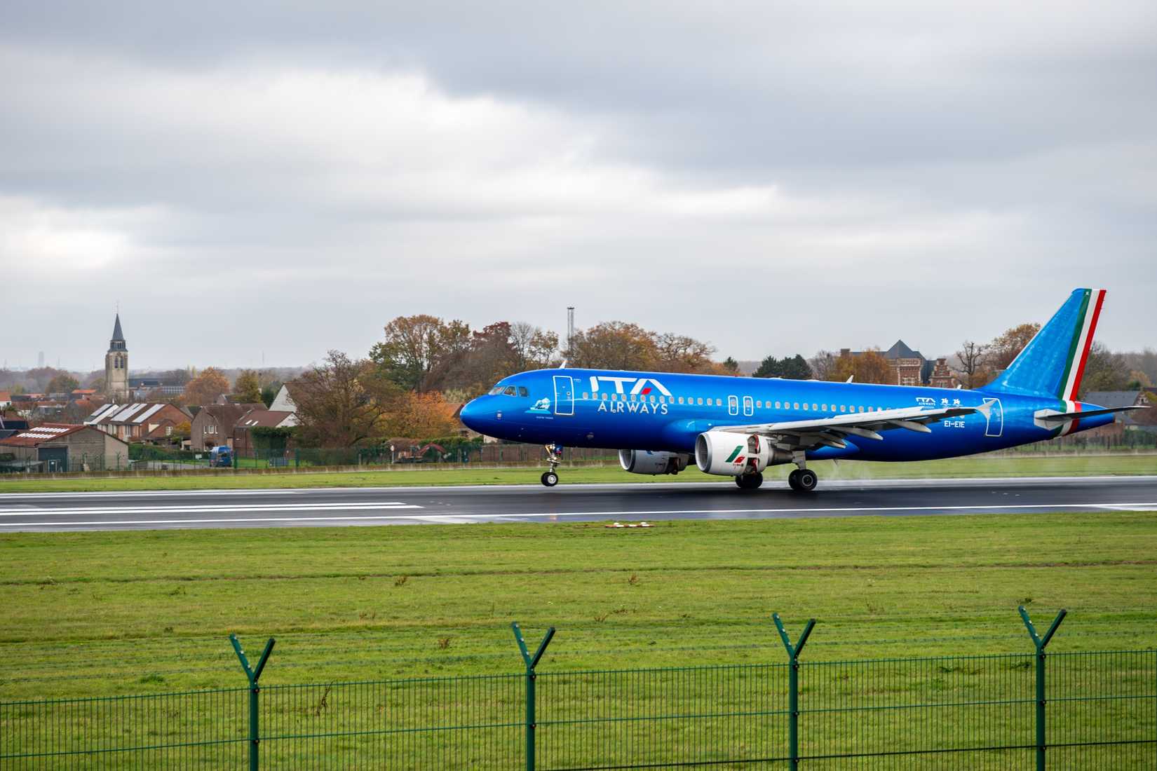 ITA Airways jet landing on the runway at Brussels Airport (Zaventem) on a wet, overcast day.