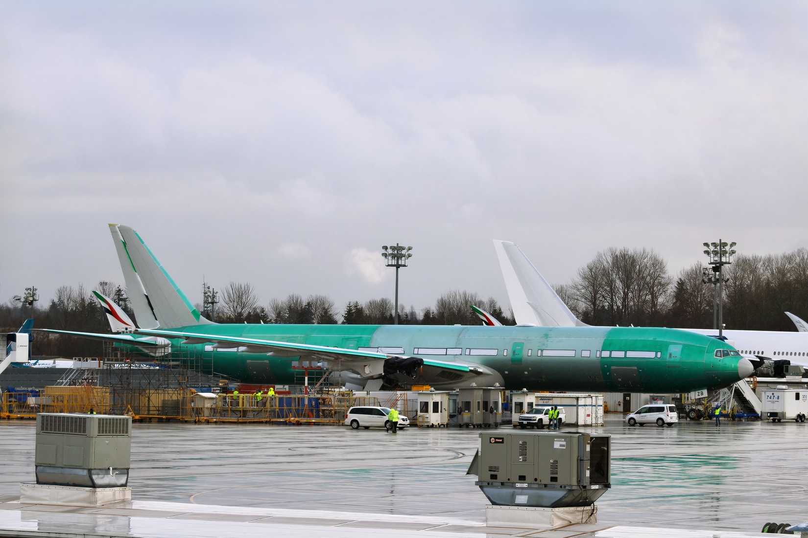 A large, wide-body aircraft, a Boeing 777X, sits on a wet tarmac under a gray, overcast sky. The aircraft is painted in a distinctive teal-green prototype color but features the red, green, and white Emirates flag striping visible near the base of its vertical stabilizer (tail fin). The scene shows ground crew, service vehicles, and scaffolding around the fuselage, with the tail of another large white jet visible in the distance.