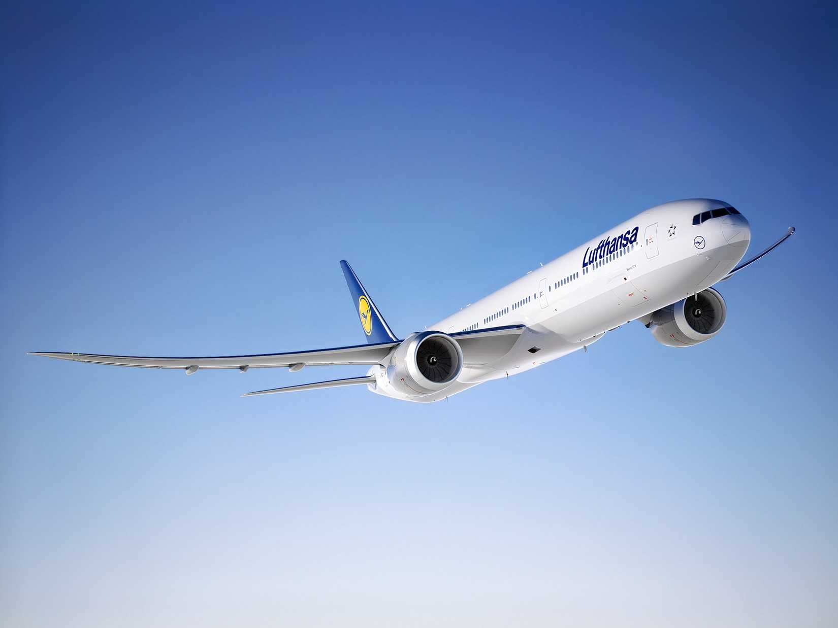 A low-angle, three-quarter view of a Lufthansa Boeing 777X passenger jet ascending against a clear, gradient blue sky. The aircraft is painted in the airline's blue and white livery, featuring the 'Lufthansa' title on the fuselage and the prominent crane logo on the blue vertical stabilizer. The image emphasizes the aircraft's large size and distinctive long wingspan.
