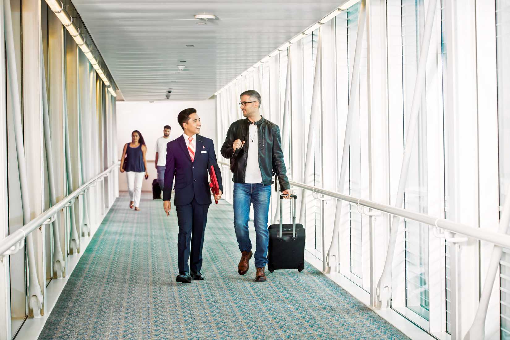  An airport service representative, wearing a dark suit and red tie, escorts a male passenger wearing a black leather jacket and jeans through a brightly lit airport jet bridge. The passenger is pulling a black rolling suitcase and is engaged in conversation with the representative. The walkway features a distinctive turquoise patterned carpet and tall windows with a white structural frame. Two other travelers are visible in the background.