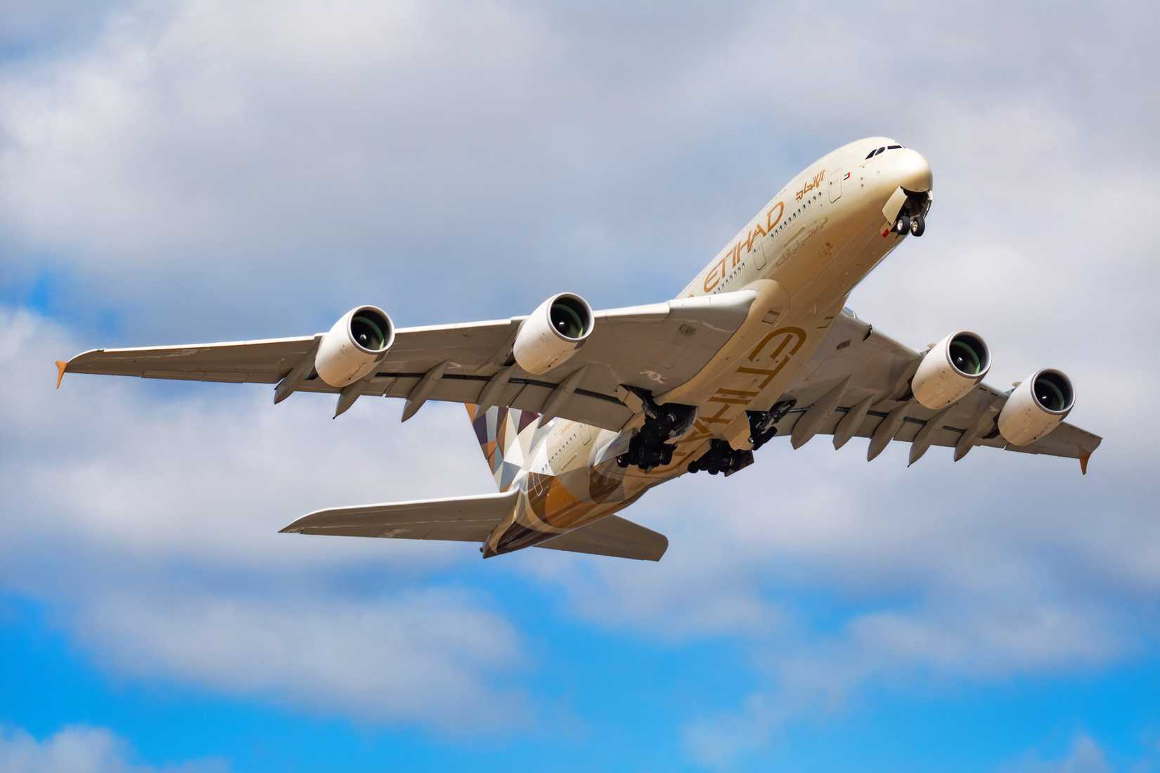 A low-angle shot of a massive Etihad Airways Airbus A380 in mid-air, ascending steeply after takeoff against a backdrop of white clouds and blue sky. The aircraft's landing gear is extended and in the process of retracting. The plane's livery is a light beige-gold, with the word "ETIHAD" in large gold letters on its underbelly and the airline's geometric tail fin design visible.