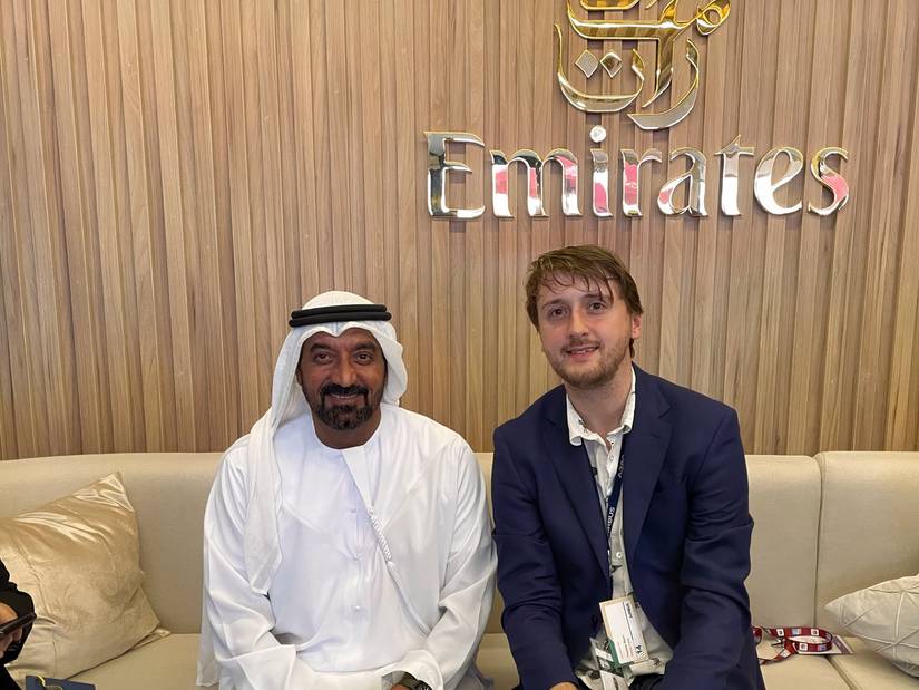 Two men are seated side-by-side on a light-colored sofa. On the left, Sheikh Ahmed is wearing traditional white Emirati attire. On the right, Tom Boon is wearing a navy blue blazer over a white shirt. They are smiling at the camera. The background is a light wood paneled wall featuring large, metallic, three-dimensional "Emirates" branding in both English and Arabic calligraphy.