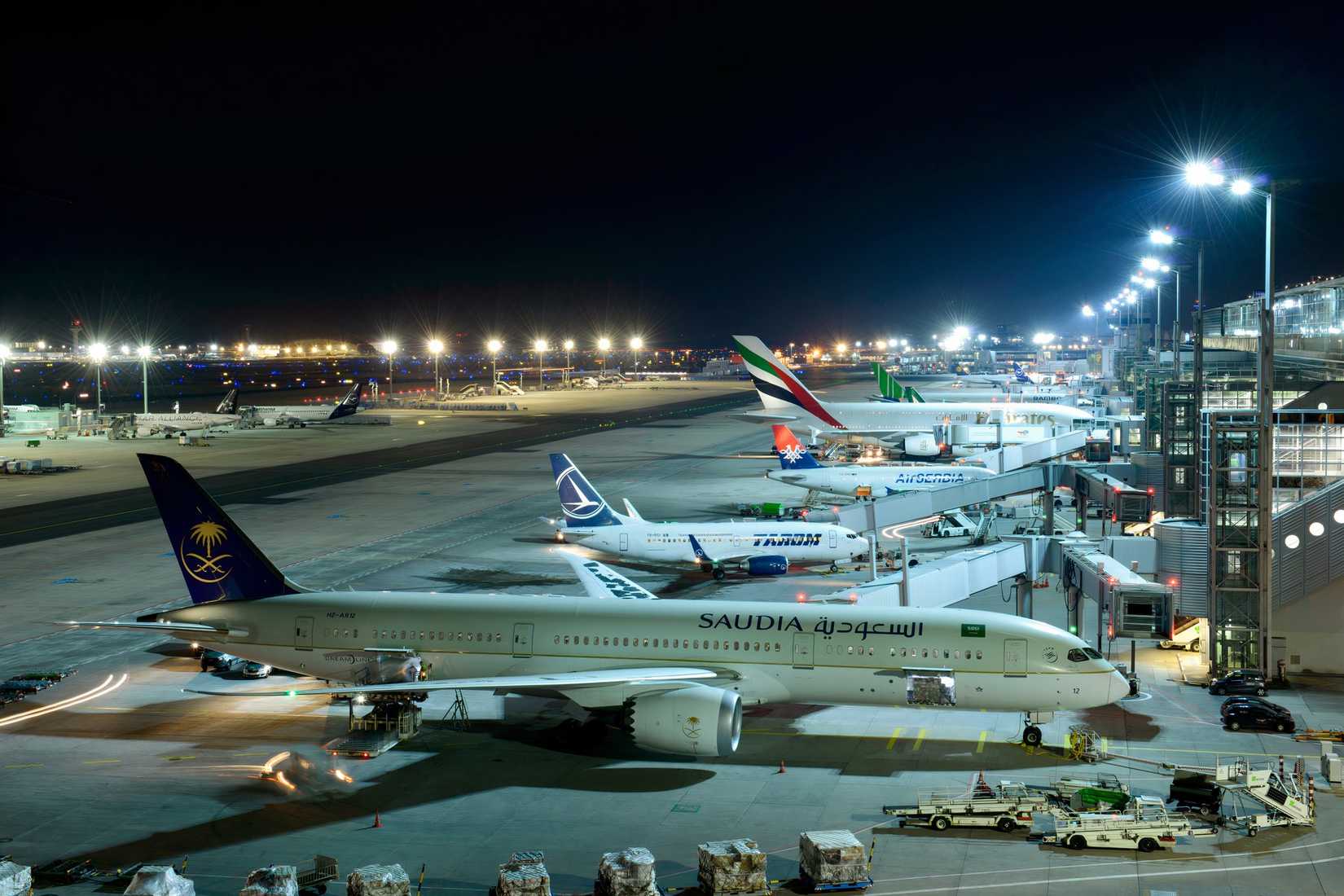 A close-up photograph of a Saudia (Saudi Arabian Airlines) Boeing 787-9 Dreamliner, registration HZ-AR12, parked at a Frankfurt Airport gate at night. The aircraft is being serviced by ground crew and a cargo loader. Several other commercial airplanes are parked at adjacent gates, including a Tarom Boeing 737, an Air Serbia aircraft, and an Emirates Boeing 777. The terminal building is brightly illuminated with multiple spotlights, and the background shows the airport's tarmac lights stretching 