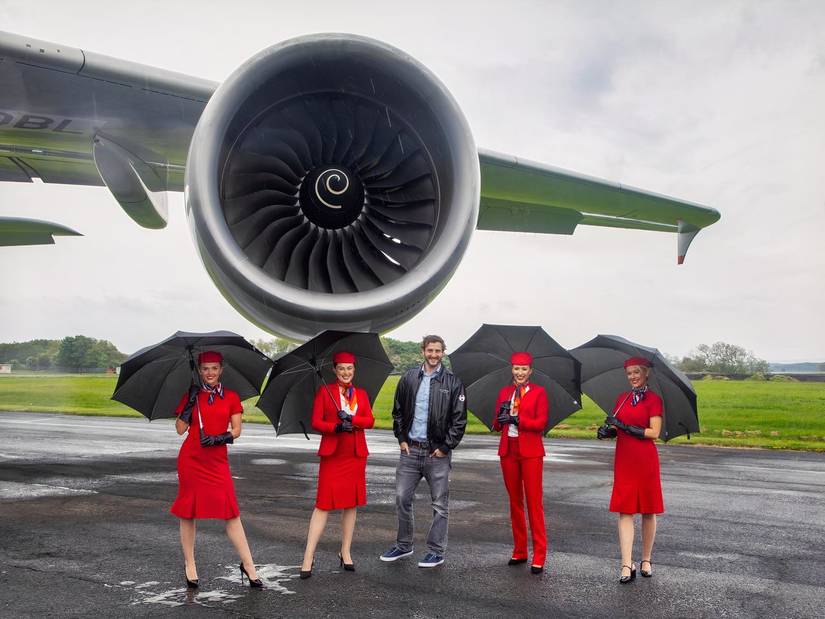 Four women in striking red flight attendant uniforms and hats, holding black umbrellas, pose with a casually dressed man beneath the massive engine and wing of a large jet aircraft. The group is standing on a wet tarmac against a backdrop of green grass under an overcast sky.