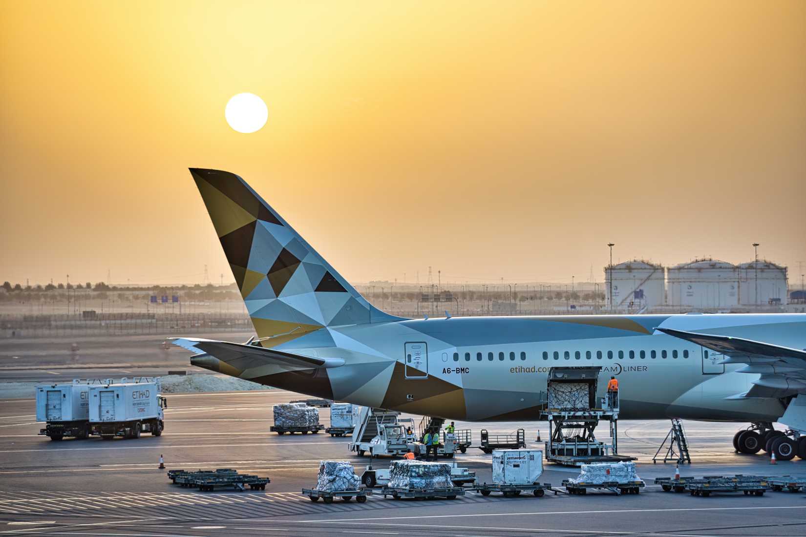 Etihad Airways Boeing 787 Dreamliner Loading or Unloading Cargo During Morning Orange Sunrise Over Tail of Plane. 