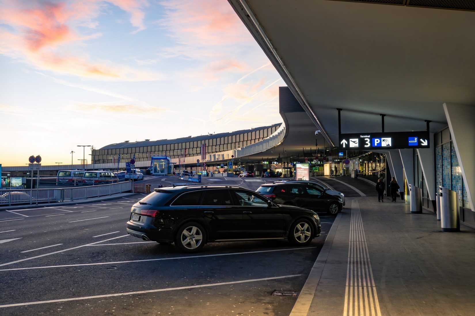An wide-angle photograph of an airport terminal exterior at dawn or dusk, with the sky showing soft pink and orange clouds. In the foreground, a black car is parked in a designated drop-off zone under a large concrete overhang. Directional signage for "Terminal 3" and parking is visible above a sidewalk where a few people are walking. In the background, the modern, curved glass facade of the airport building stretches across the horizon.