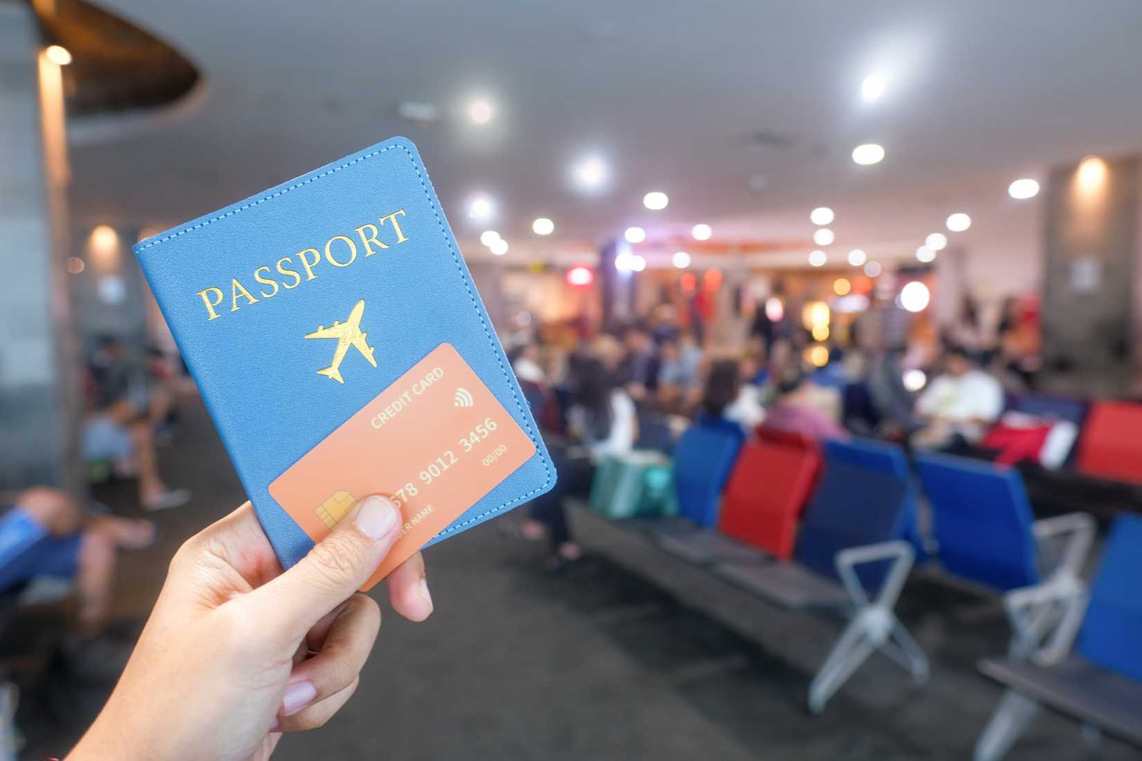 A person holds a blue passport wallet and a credit card in a busy airport lounge, with waiting passengers in the background