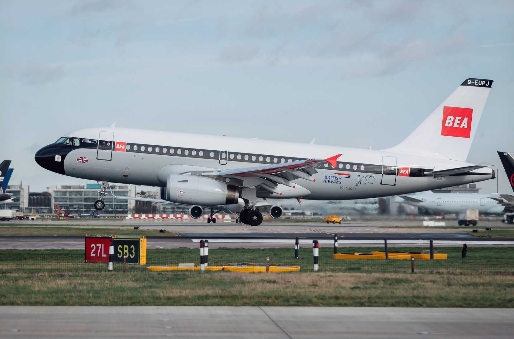 A British Airways Airbus A319-100, featuring a retro British European Airways (BEA) "Red Square" heritage livery and registration G-EUPJ, is captured just after landing on a runway. The aircraft has a black nose, a grey upper fuselage with a white roof, and a large red square logo with white "BEA" text on the tail. It is seen in a side profile on the airfield with its landing gear down, and airport signage for runway 27L is visible in the foreground under an overcast sky.