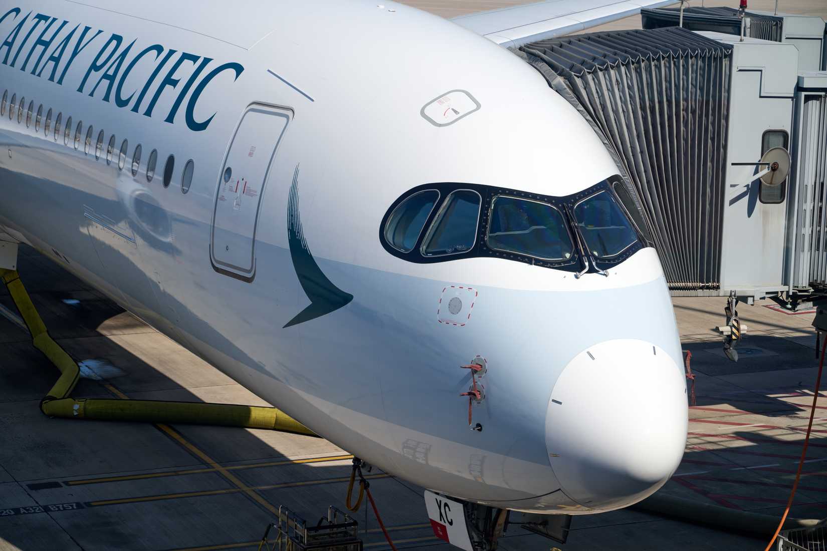 Close-up of Cathay Pacific A350 nose and cockpit at HKG gate, jet bridge attached under sunny skies.