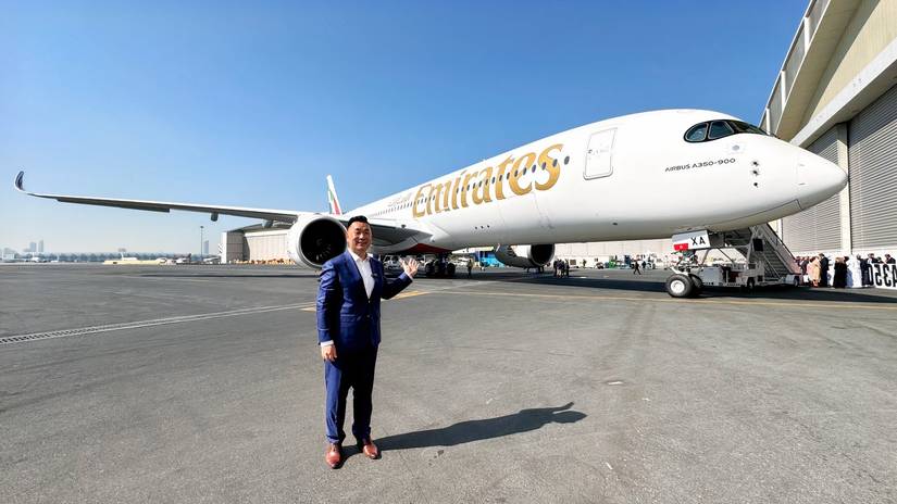 Sam Chui in a blue suit stands on a sunny tarmac, gesturing toward a large Emirates Airbus A350-900 parked behind him. The aircraft features the gold "Emirates" title on its white fuselage and the UAE flag on its tail. A mobile boarding stair is connected to the front door, and a large hangar and other airport buildings are visible in the background under a clear blue sky.