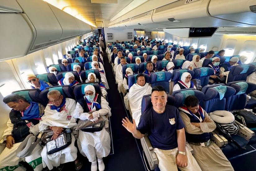 An interior view of a crowded airplane cabin, specifically a Garuda Indonesia Boeing 747 configured for Hajj pilgrimage. The economy class seats are filled with passengers, many of whom are wearing traditional white pilgrimage attire (Ihram) or head coverings. In the foreground, Sam Chui in a dark t-shirt with a small Shiba Inu logo sits in an aisle seat, waving at the camera. The cabin features blue seating with teal headrest covers, and overhead bins line both sides of the center aisle.