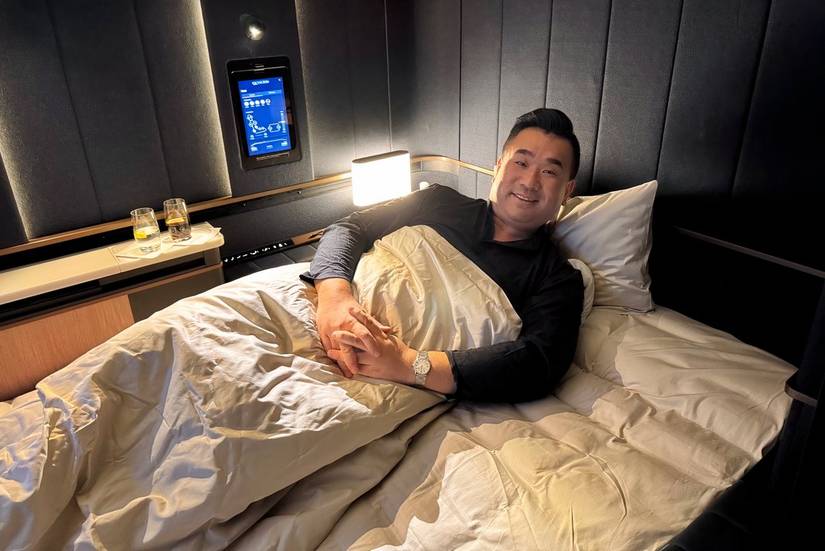 Sam Chui lies comfortably in a large, fully flat bed within a luxury aircraft cabin, the Lufthansa Allegris First Class suite. He is tucked under a white duvet with his hands clasped, smiling at the camera. The suite features dark, padded walls, a personal touch-screen controller mounted on the wall, a small bedside lamp, and two glasses of water on a side console. The lighting is warm and subdued, creating a cozy, private atmosphere.