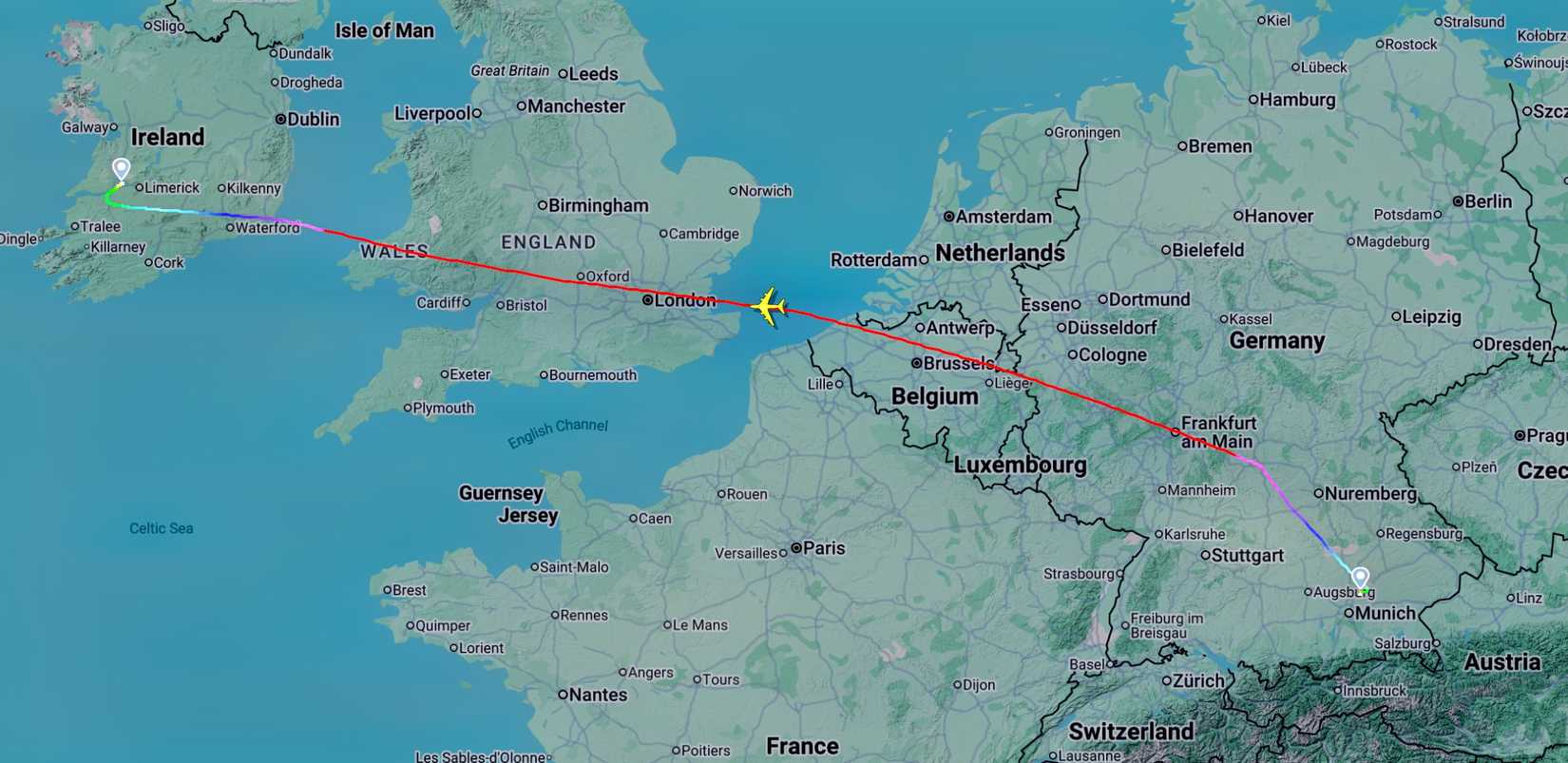 A digital map of Western Europe showing a flight path tracking to Ireland from Southern Germany. The path ends near Limerick, Ireland, indicated by a blue pin, and travels southeast across the Irish Sea, Wales, and Southern England. The line continues over the English Channel, Belgium, and Luxembourg, starting at another blue pin in Augsburg, near Munich, Germany. The flight path is represented by a multi-colored line that transitions from green to blue, pink, red, and purple, with a small yello