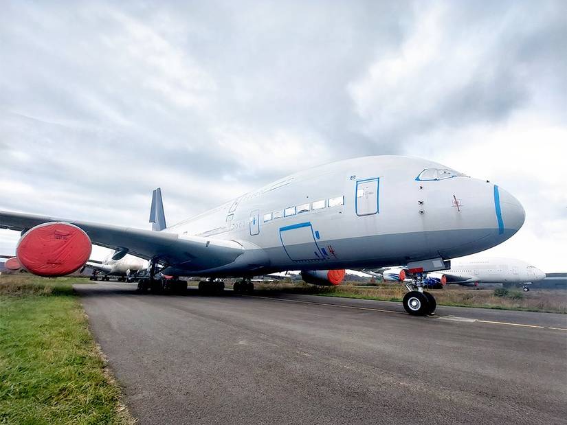 A ground-level, wide-angle shot of a decommissioned Airbus A380 parked on a paved storage area under a thick, grey overcast sky. The massive aircraft is painted in a plain white and grey livery with its windows, doors, and various sensors sealed with blue and silver protective tape. Its large engines are covered with bright red protective caps. The plane is parked on an asphalt surface bordered by green grass, with at least two other large wide-body aircraft visible in the background, also in st