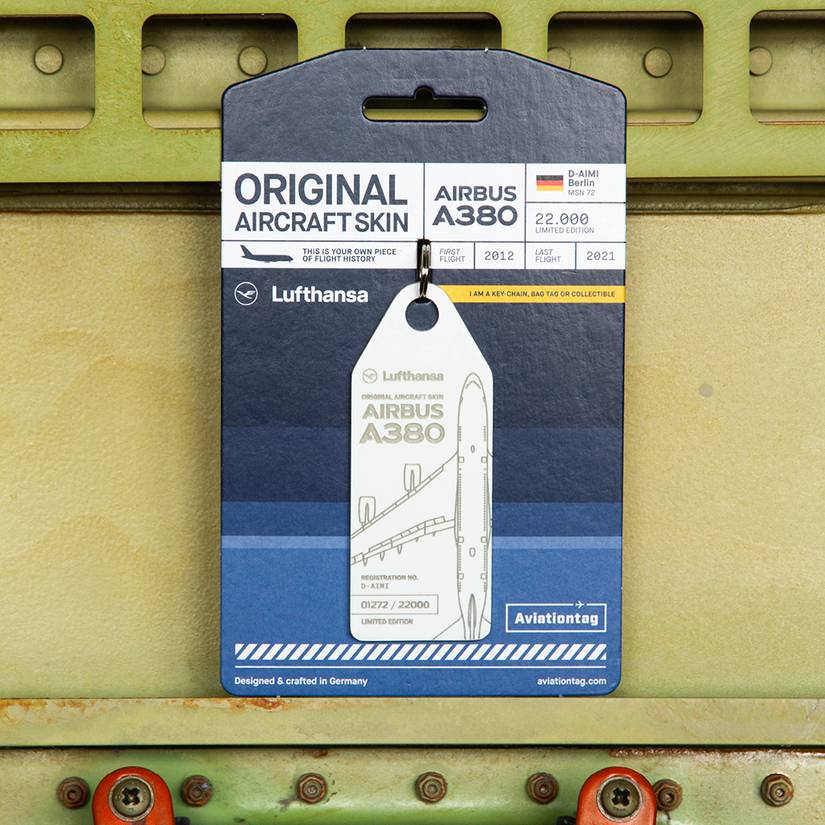 A white aircraft skin tag is attached to a blue and white display card with a black hanging slot at the top. The card reads "ORIGINAL AIRCRAFT SKIN" and "AIRBUS A380," featuring the Lufthansa logo and details for aircraft D-AIMI (MSN 72), noting a first flight in 2012 and a last flight in 2021. The tag itself is white with a gray engraved outline of an A380 and the text "AIRBUS A380" and "REGISTRATION NO. D-AIMI." The entire item is photographed against a background of light green industrial met