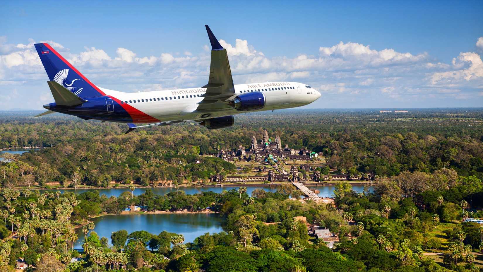 An Air Cambodia Boeing 737 MAX is captured in flight, banking to the right over a lush tropical landscape. The aircraft features a white fuselage with "AIR CAMBODIA" titles and a navy blue tail decorated with a white stylized bird logo and the Cambodian flag. In the background, the ancient stone spires of Angkor Wat rise from the dense green forest, surrounded by a large moat and reflecting pools under a clear blue sky with scattered white clouds.
