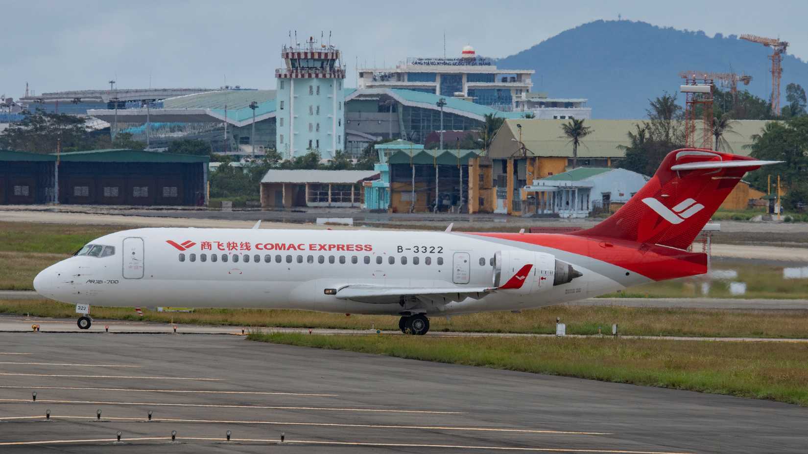 A COMAC ARJ21-700 aircraft with the registration B-3322 is taxiing on an airport runway. The plane features a white fuselage with "COMAC EXPRESS" branding and a red logo, paired with a bright red tail and engine cowlings. In the background, airport facilities including a control tower, hangars, and construction cranes are visible under an overcast sky.