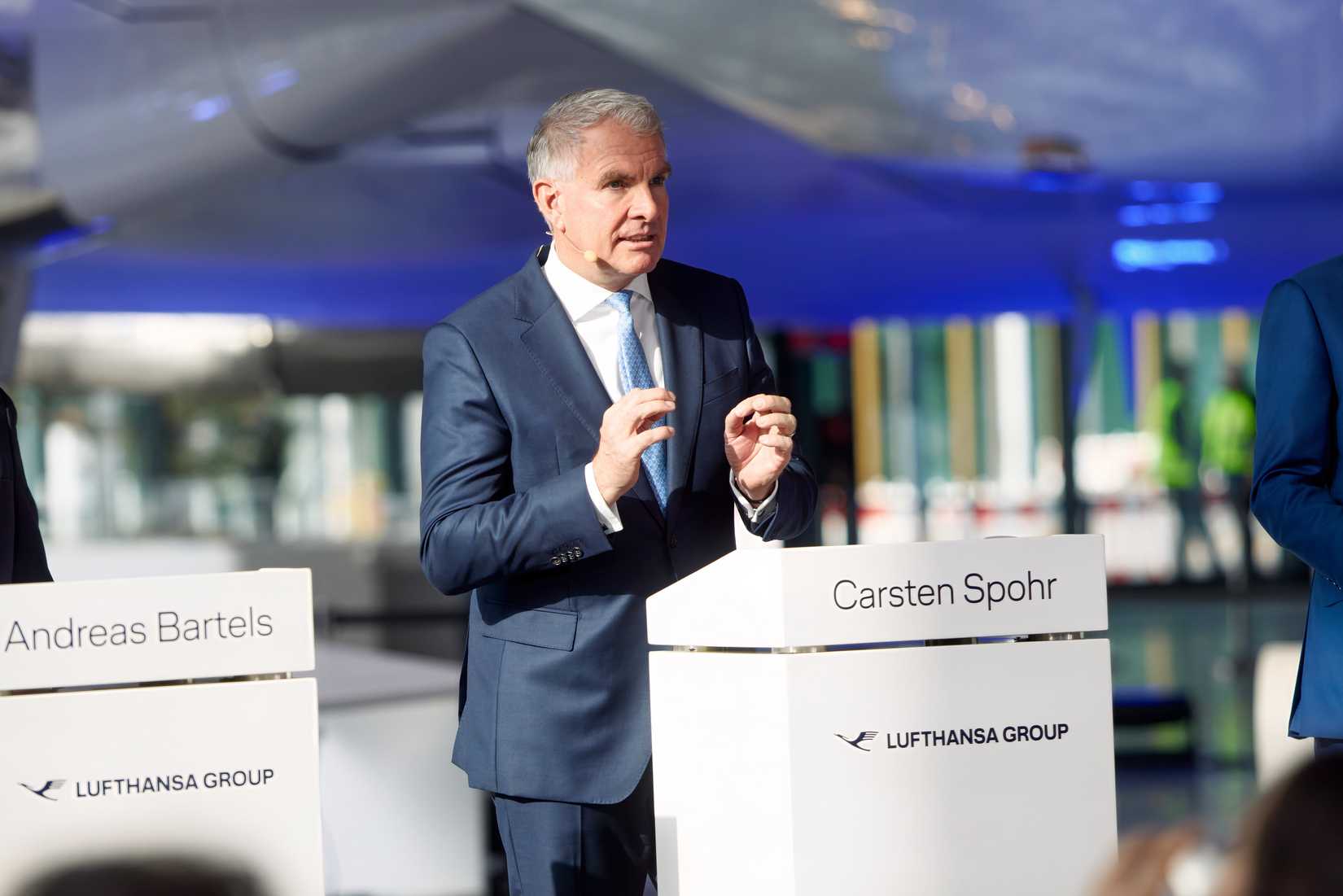 Lufthansa Group CEO Carsten Spohr speaks at a podium during a formal presentation, with fellow speaker Andreas Bartels visible at an adjacent podium, set against a backdrop featuring a large aircraft fuselage.