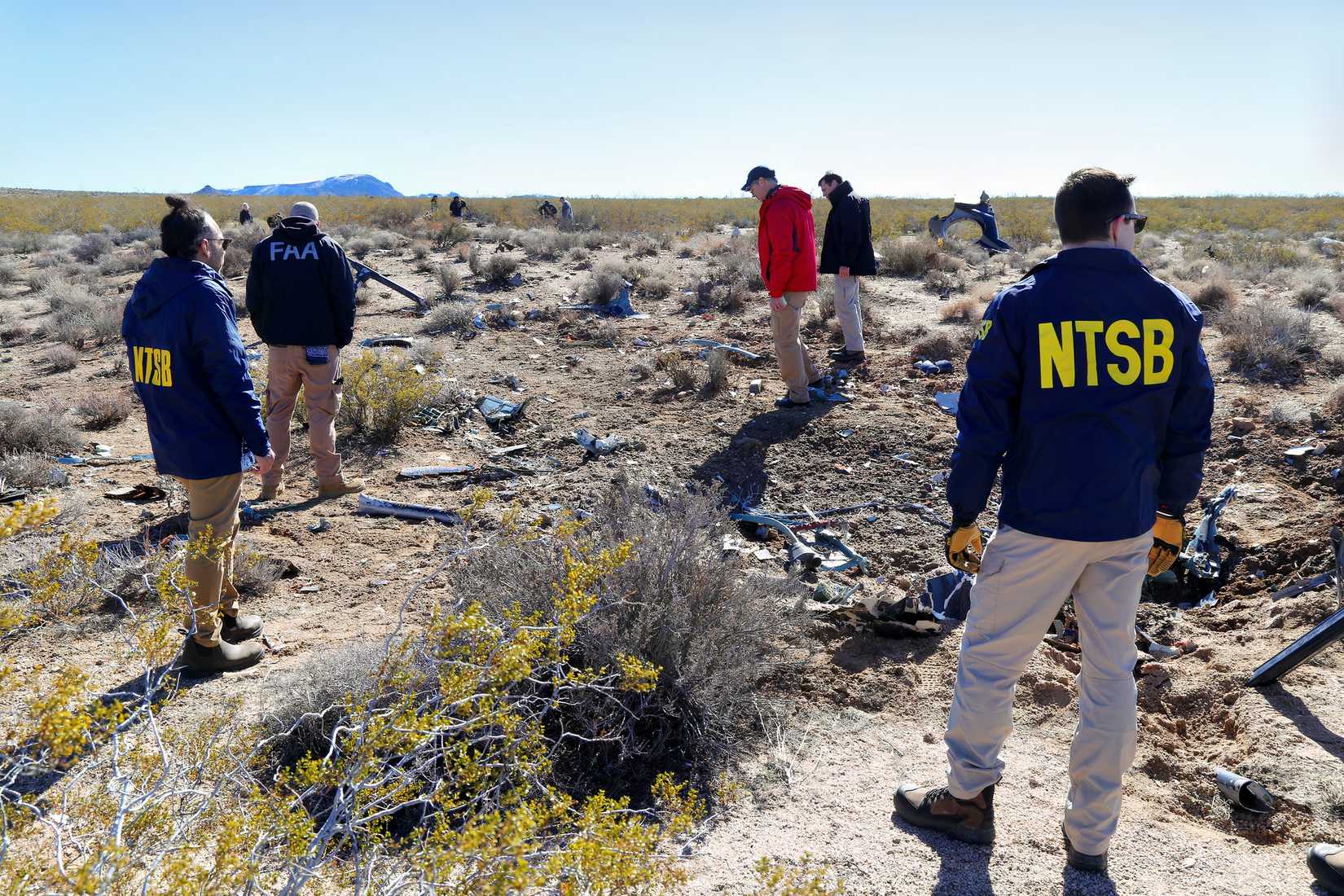 Personnel from the National Transportation Safety Board (NTSB) and the Federal Aviation Administration (FAA) are seen at a debris field in a desert landscape. Several investigators, identified by the yellow letters on their navy blue jackets, are examining scattered wreckage across the arid, scrub-covered ground under a clear sky. In the background, other members of the investigation team are spread out across the site, with distant mountains visible on the horizon.