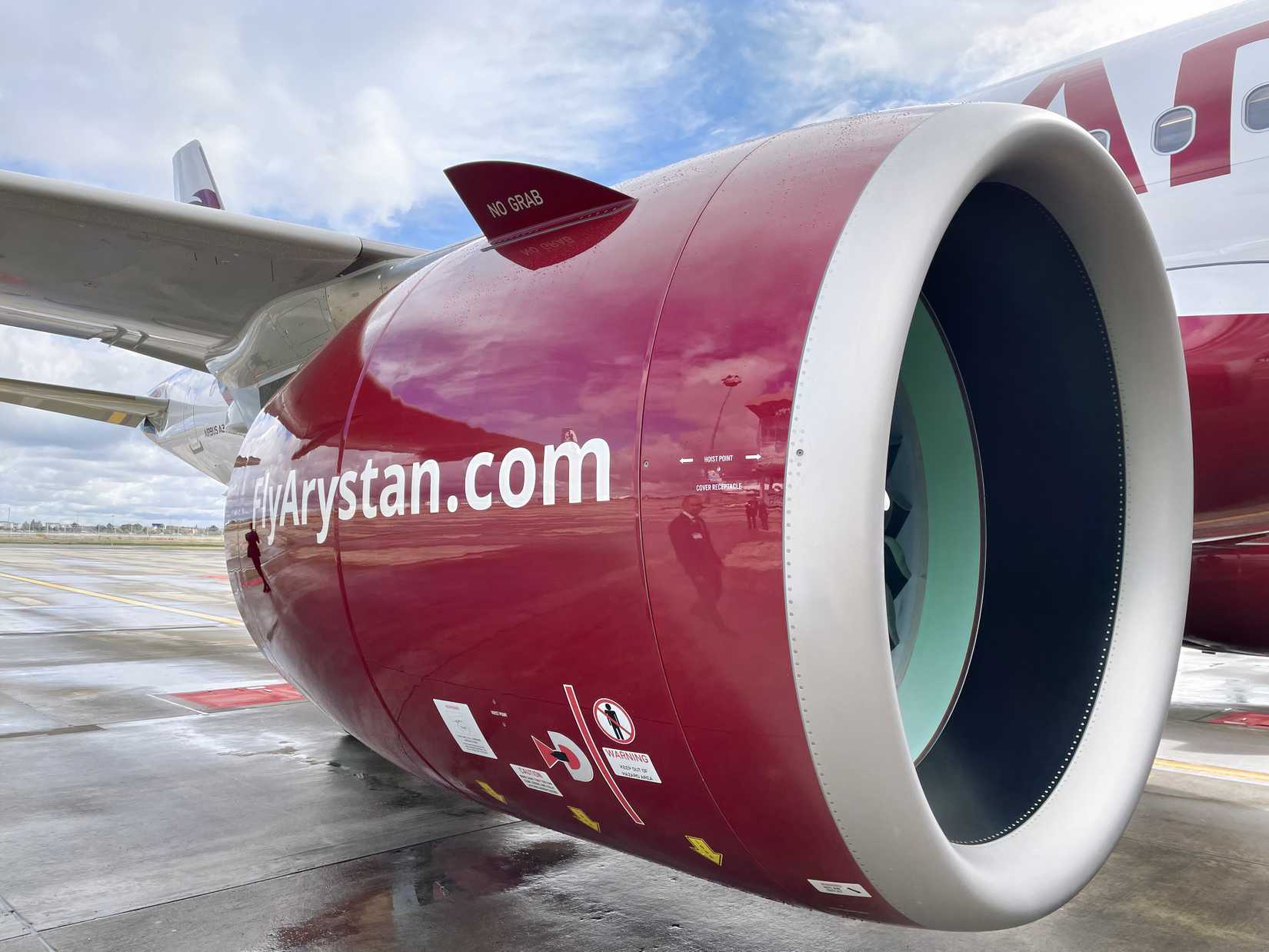 A close-up side view of a large aircraft engine cowling on a FlyArystan plane, painted deep red with "FlyArystan.com" in white text. The engine intake is prominent in the foreground, showing the silver rim and the interior fan blades. A small aerodynamic fin on the upper side of the cowling is labeled "NO GRAB," and various technical warning labels are visible near the bottom. The wet tarmac and a cloudy sky are visible in the background.