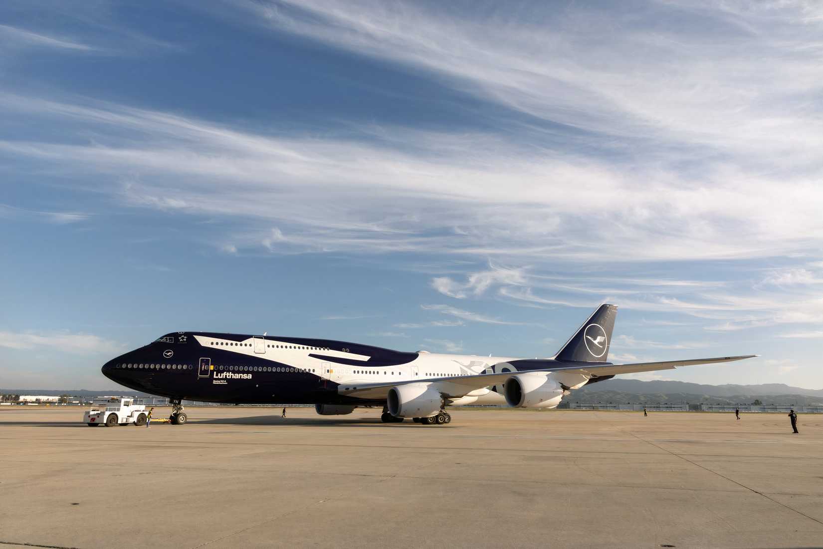 A Lufthansa Boeing 747-8 in the airline's dark navy blue livery being towed across an airport apron by a pushback tug, set against a blue sky with wispy clouds and mountains in the background.
