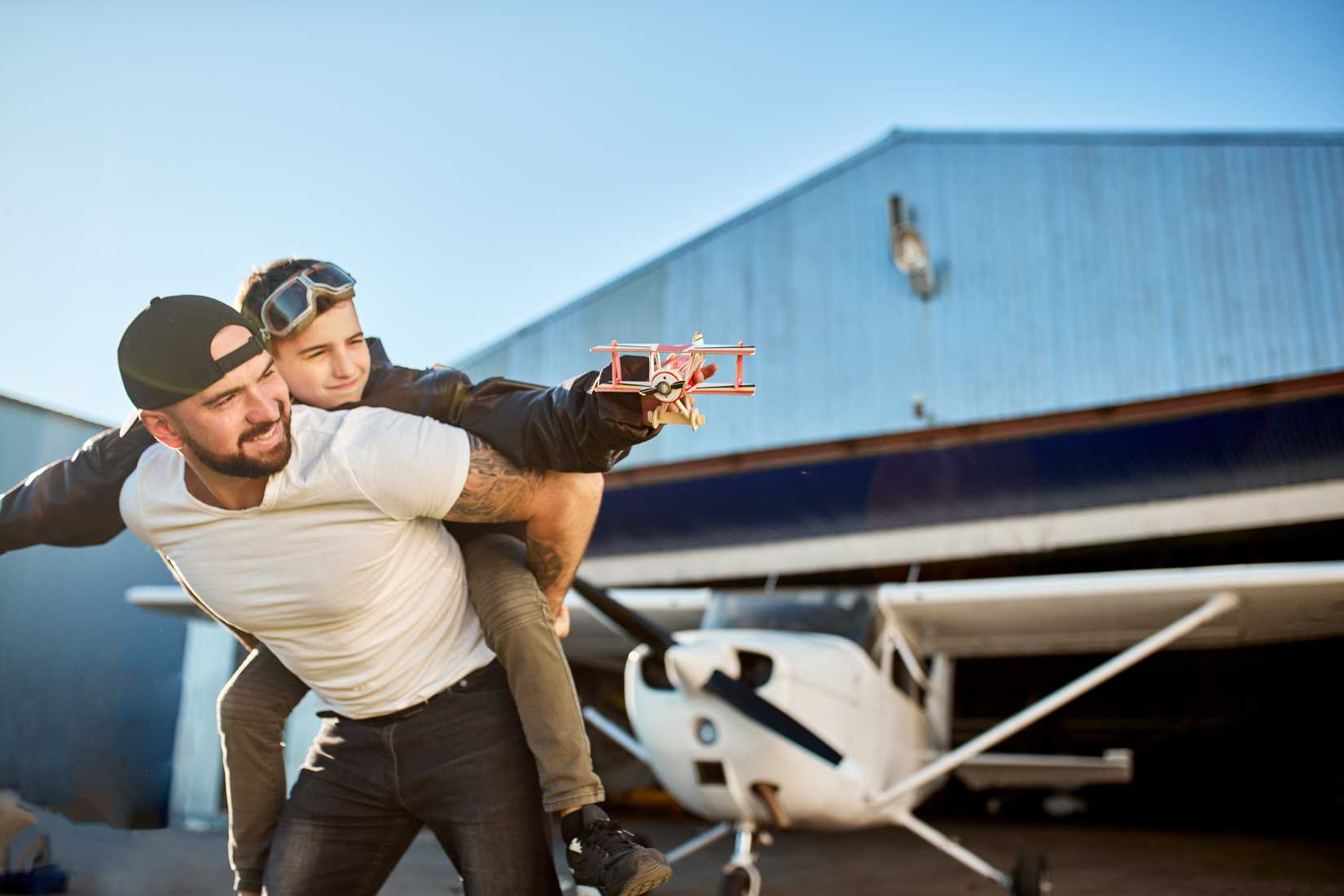 A young dad holding on his back little son in aviator glasses and large pilot jacket, holding handmade toy plane, playing outside aircraft shed, smiling and feeling happy.