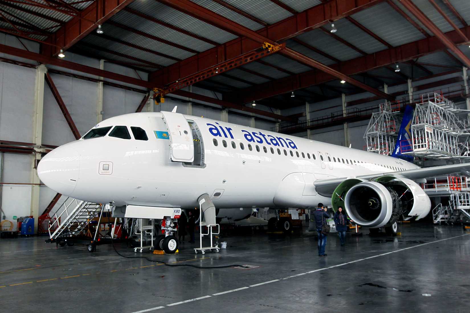 An Air Astana Airbus A321neo is shown inside a large maintenance hangar with its passenger door open and a staircase attached. The aircraft's left engine cowling is open, revealing the complex internal components for maintenance or inspection. Two people are standing on the hangar floor near the engine, and various scaffolding and equipment are visible in the background under the industrial metal roof.