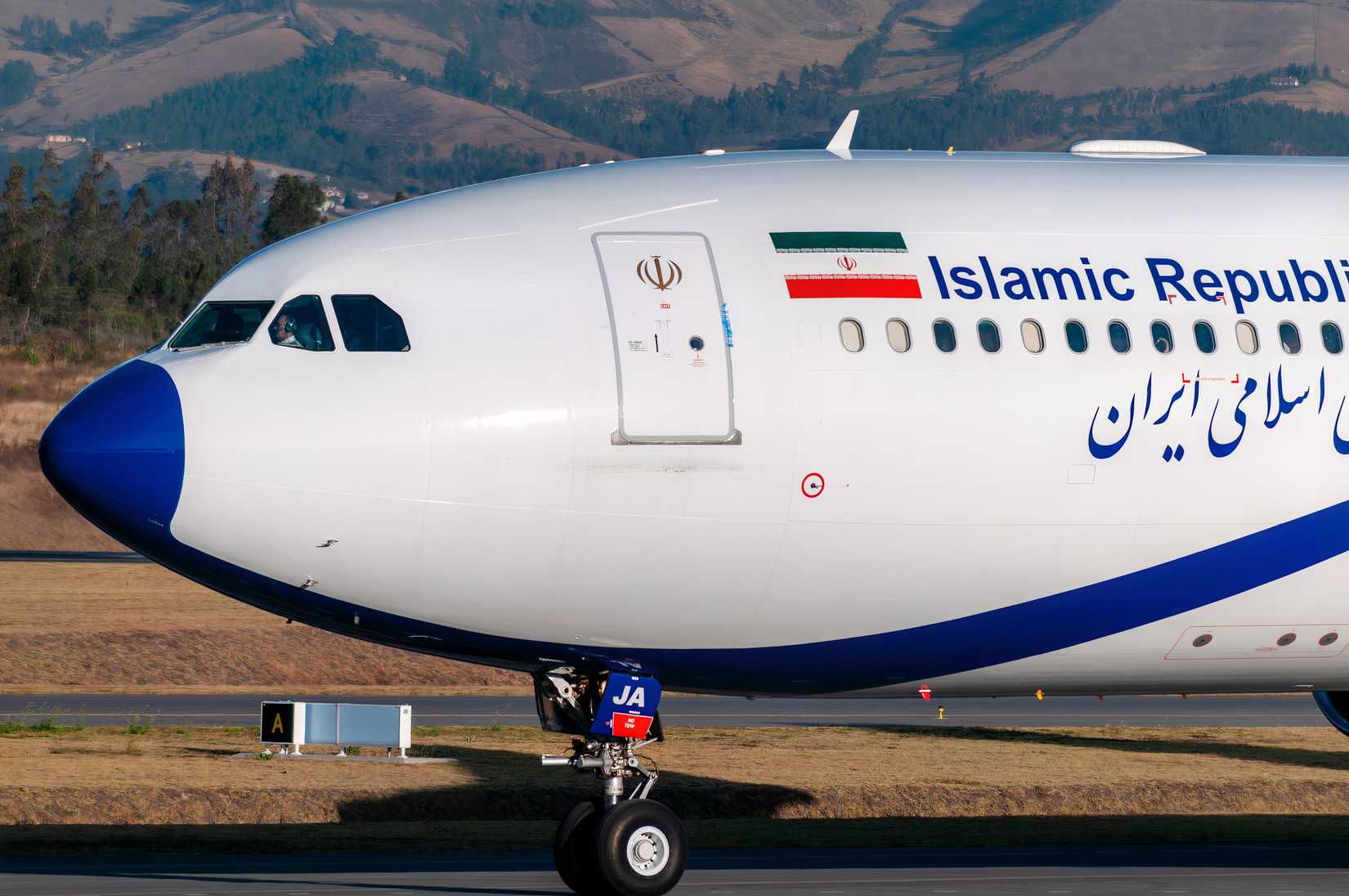 A close-up shot of the nose and cockpit area of an Airbus A340-300 belonging to the Islamic Republic of Iran. The aircraft features a white fuselage with a prominent blue nose cone and the "Islamic Republic of Iran" titles in both English and Persian script. The Iranian flag and national emblem are visible near the forward passenger door, and a pilot can be seen through the cockpit window as the plane taxis past a grassy airfield with mountains in the background.