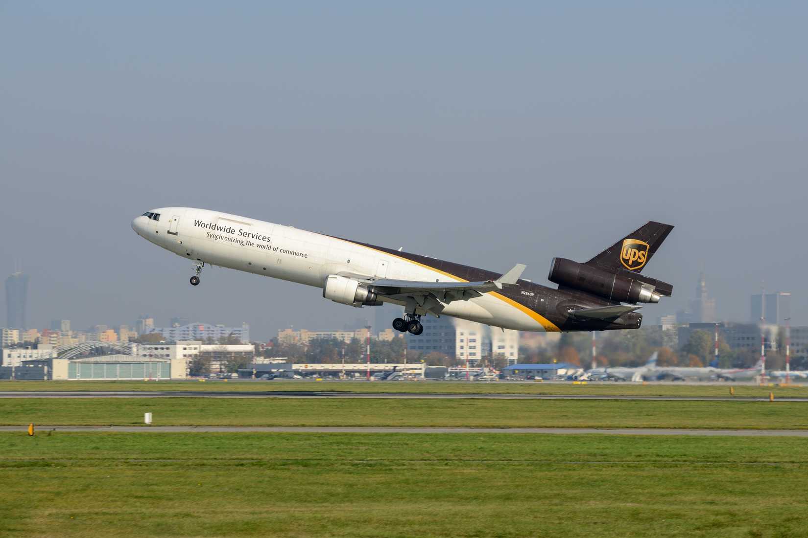 UPS MD-11 taking off surrounded by green grass and many aircraft far behind in the background