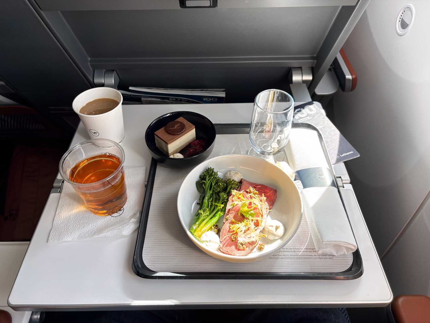 An airline meal is served on a fold-down tray table, featuring a main dish of sliced roast beef with broccolini, a chocolate layer cake for dessert, and various beverages. The service includes a Lufthansa-branded paper cup, a glass of amber-colored liquid, a small water glass, and a cloth napkin roll, all part of a special commemorative flight experience.