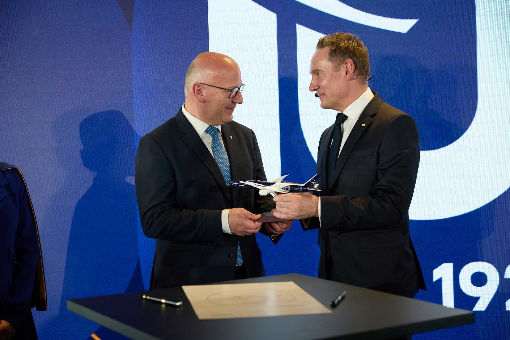 Two men in dark suits stand together in front of a blue commemorative backdrop, holding a small model of a Lufthansa aircraft featuring a special navy blue and white anniversary livery. The man on the right, Jens Ritter, is presenting the model to the man on the left, both smiling as they hold the gift over a table containing a signed document.
