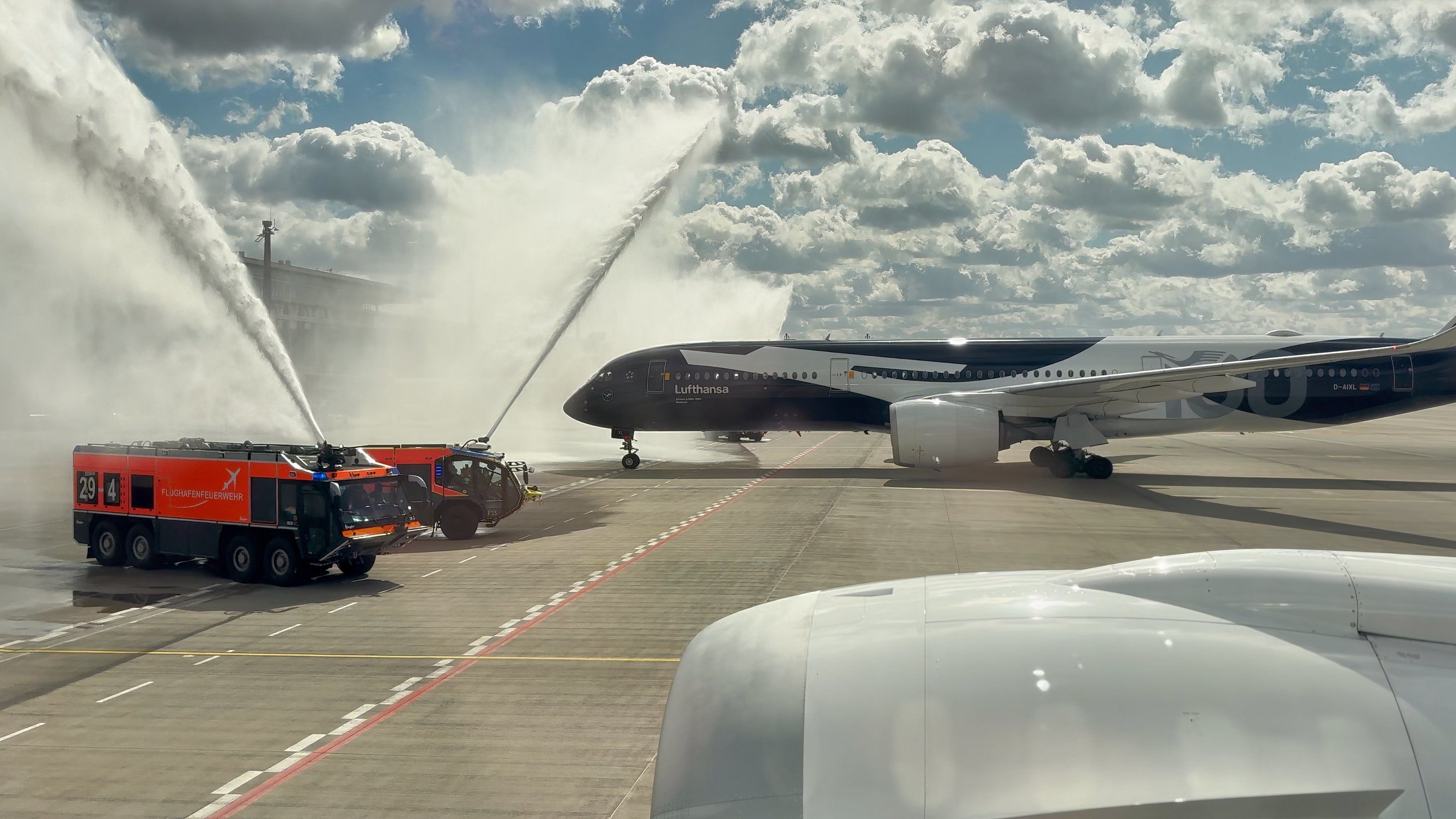 A Lufthansa Airbus A350-900 in a special navy blue and white livery receives a ceremonial water salute from two red airport fire trucks on a tarmac under a cloudy sky.