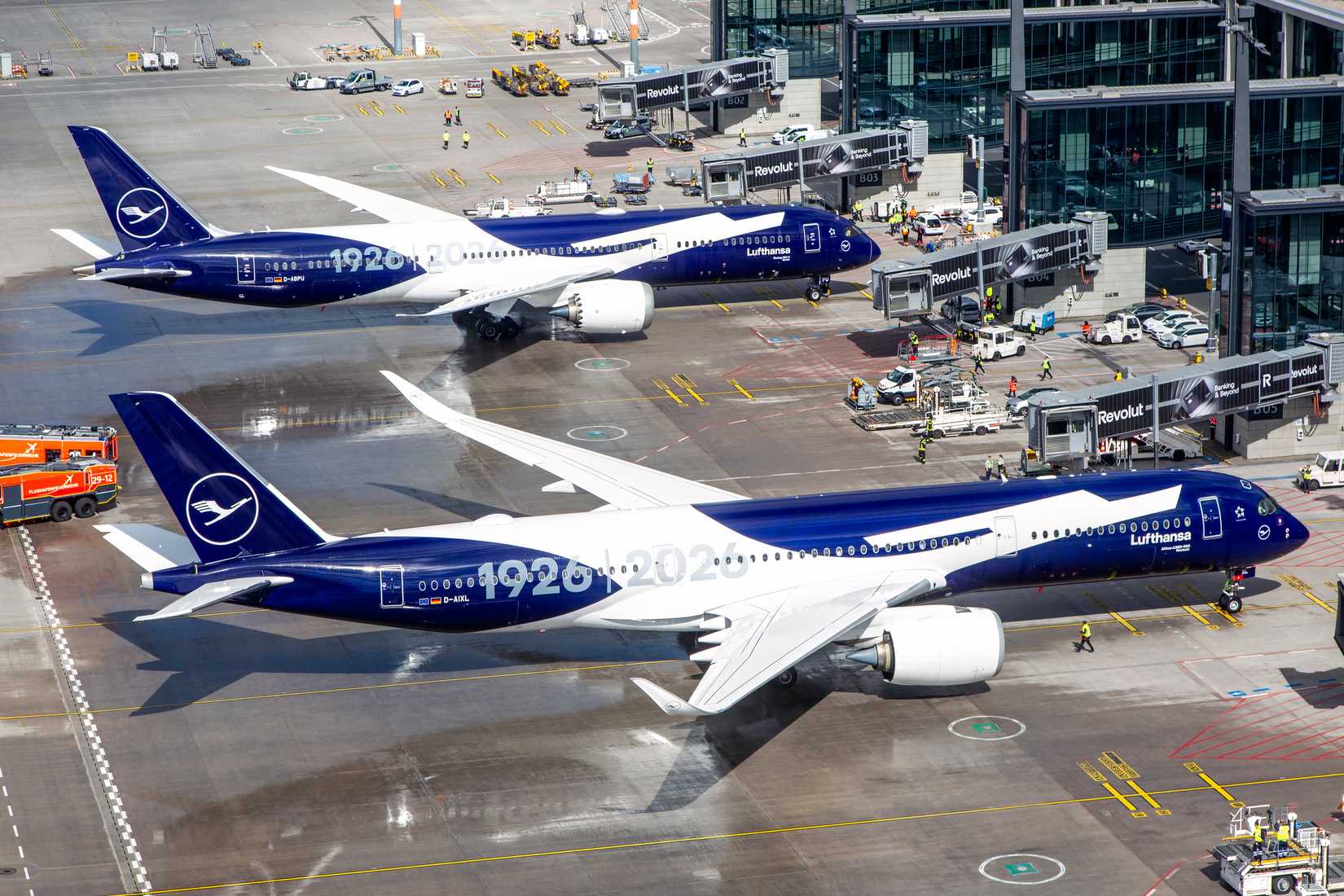 Two Lufthansa aircraft, an Airbus A350-900 and a Boeing 787-9 Dreamliner, are parked at airport gates featuring a special commemorative "1926 | 2026" livery. Both planes have a distinctive navy blue and white design with the Lufthansa logo on the tail. The scene is viewed from an elevated perspective, showing various ground service vehicles and jet bridges branded with "Revolut".