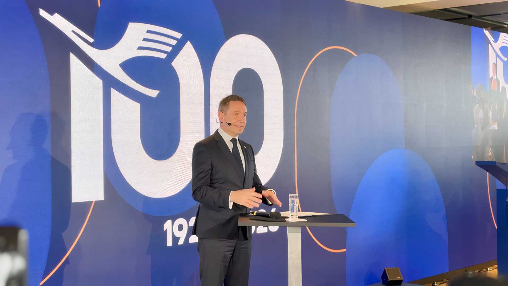 Jens Ritter, CEO of Lufthansa Airlines, speaks at a podium during an event celebrating the airline's 100th anniversary. He stands in front of a large blue backdrop featuring a stylized white "100" logo integrated with the Lufthansa crane, with the dates "1926 | 2026" visible below.