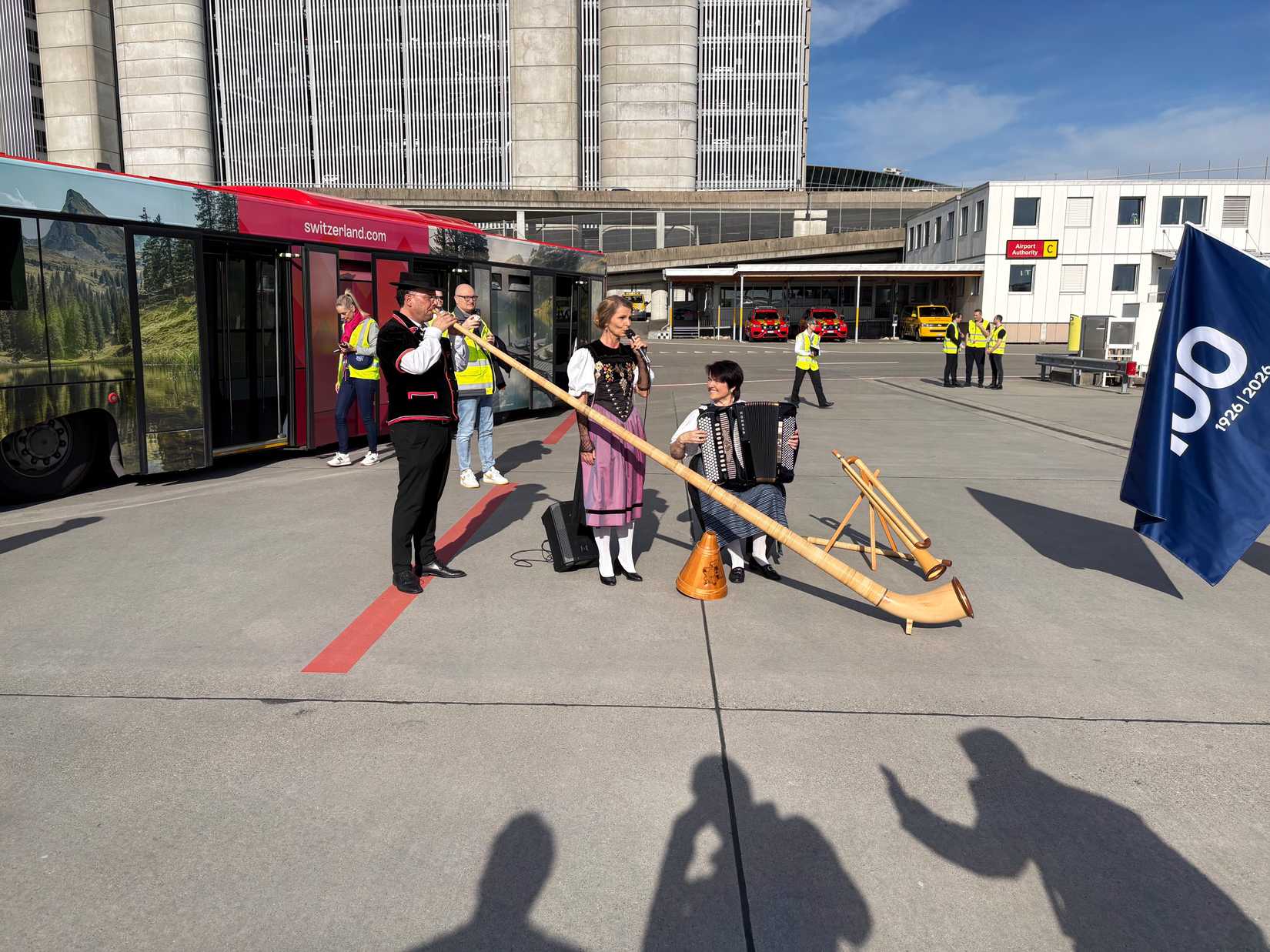 A group of performers in traditional Swiss attire perform on an airport apron next to a red bus branded with "switzerland.com." The ensemble includes a man playing a long alphorn, a woman singing into a microphone, and another woman playing an accordion, while a blue flag with a white "100" logo stands nearby.